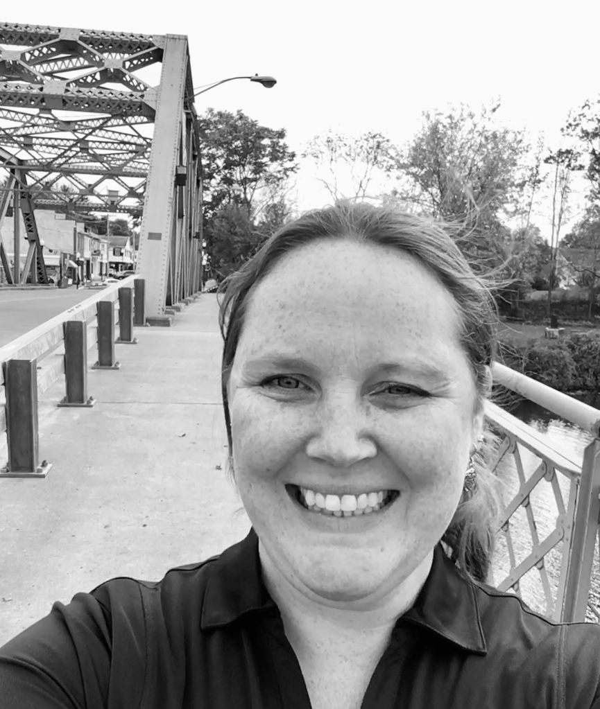 black and white image of a pale skinned person with a giant smile. They are standing on a bridge